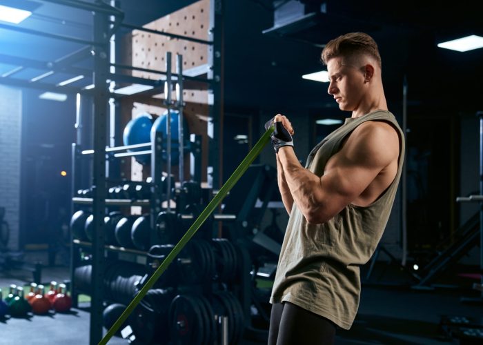 Portrait of muscular bodybuilder training arms with fitness resistance bands. Side view of man with perfect body training biceps in gym in dark atmosphere. Concept of bodybuilding.