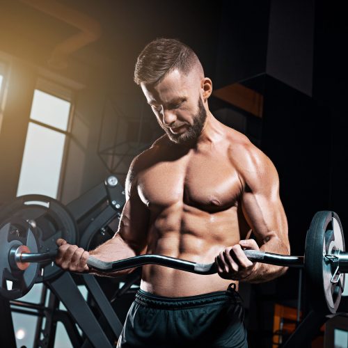 Portrait of super fit muscular young man working out in gym with barbell on gray